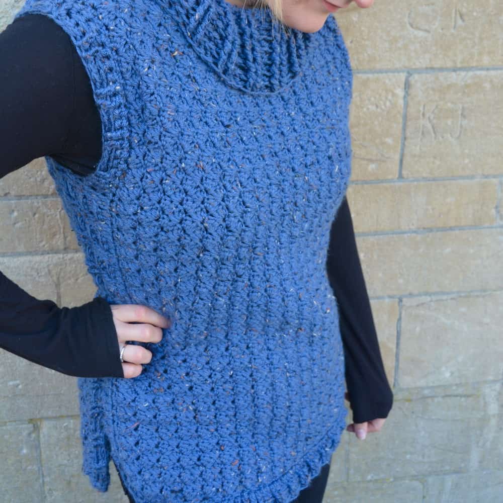 woman wearing blue tweed crochet pullover and black long sleeved top against stone wall with hand on hip