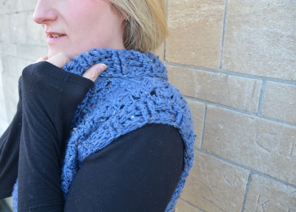 woman wearing blue tweed crochet pullover and black long sleeved top against stone wall pulling collar up