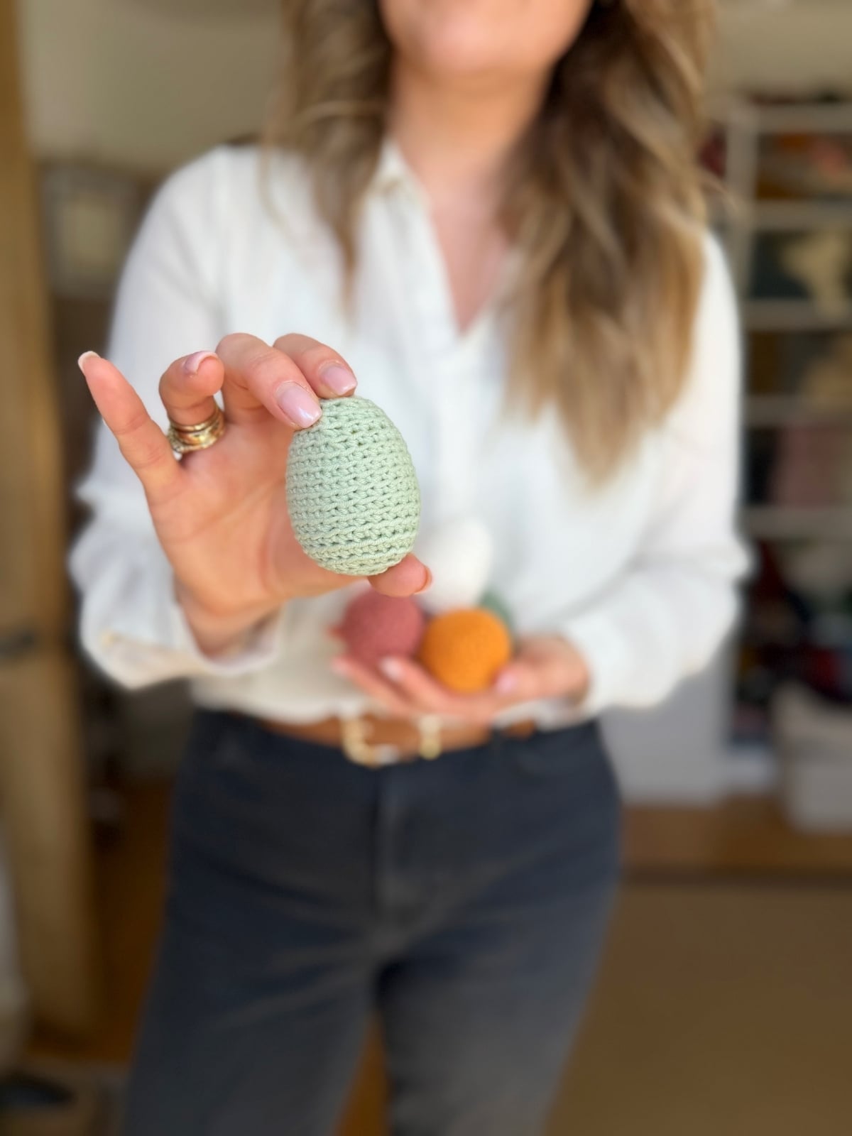 A person in a white shirt and dark pants holds a small green crochet Easter egg, with other crochet Easter eggs in their other hand.