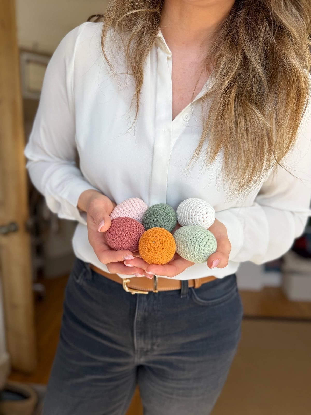 A person in a white shirt and dark pants holds six crochet Easter eggs in various pastel colors in their hands.