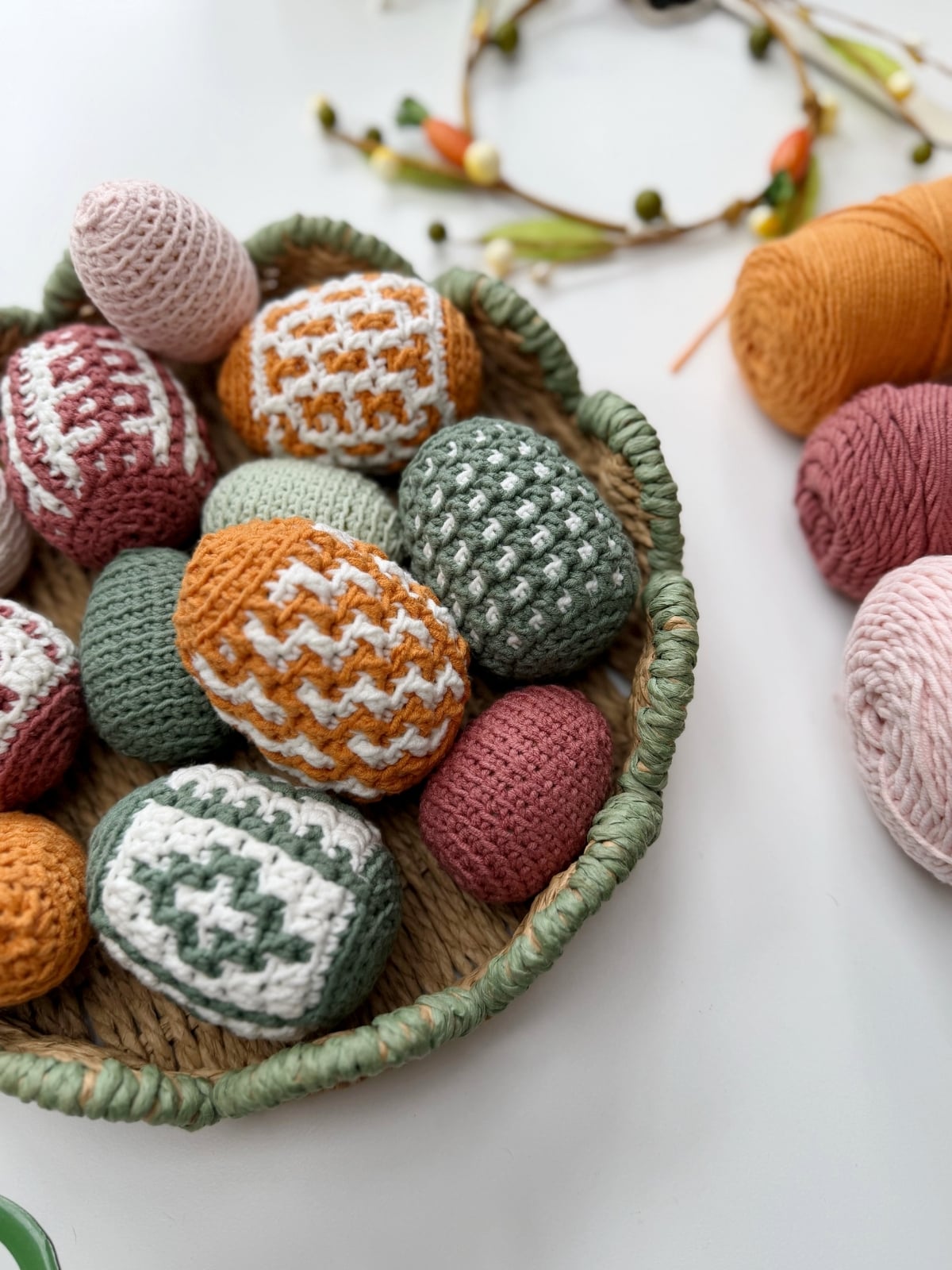 A woven basket filled with crochet Easter eggs in various patterns and colors sits on a white surface near balls of yarn and a floral decoration.
