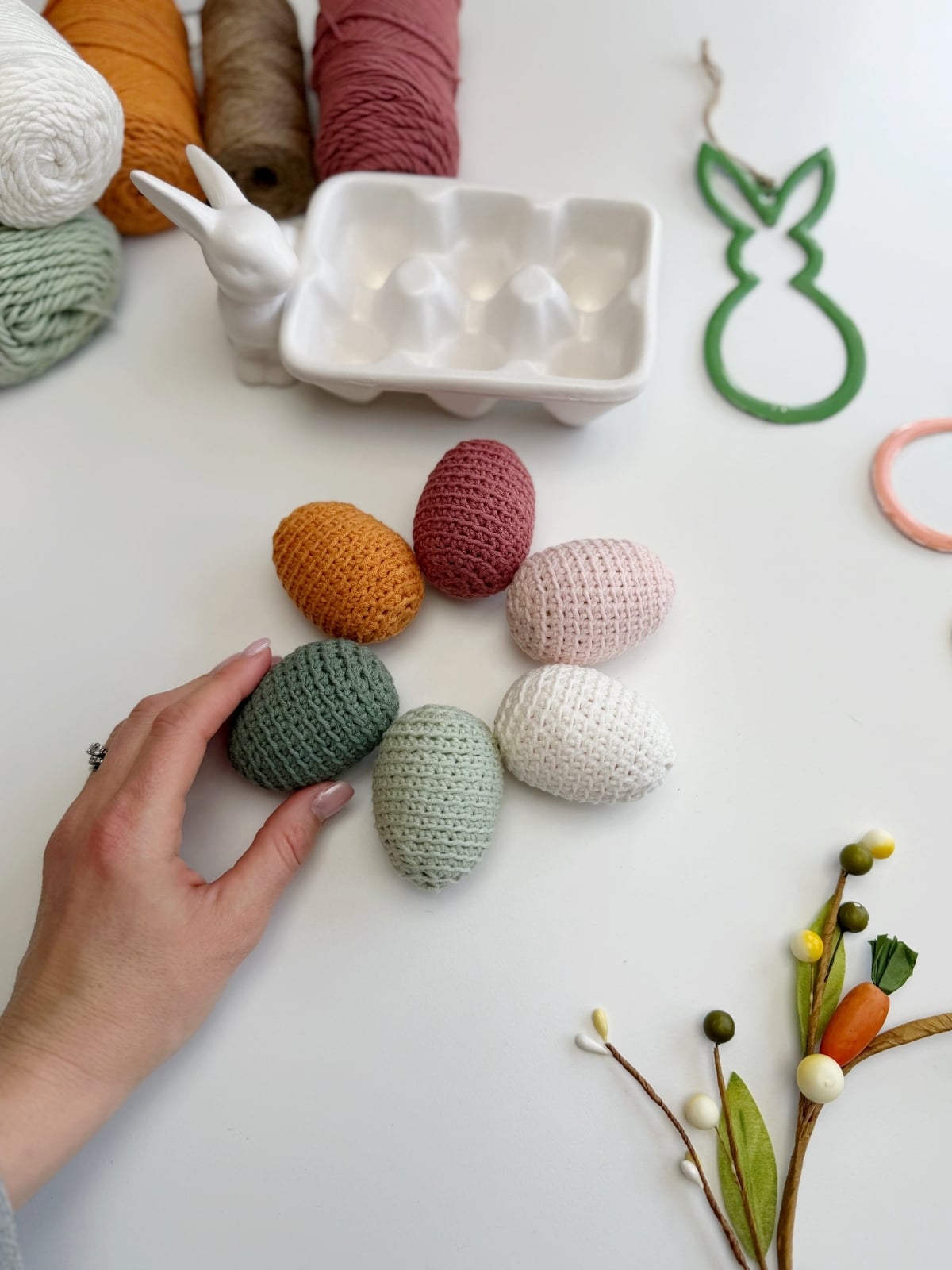 A hand touches a group of five crochet Easter eggs in pastel colors on a table with yarn, a ceramic egg holder shaped like a rabbit, and decorative craft items.