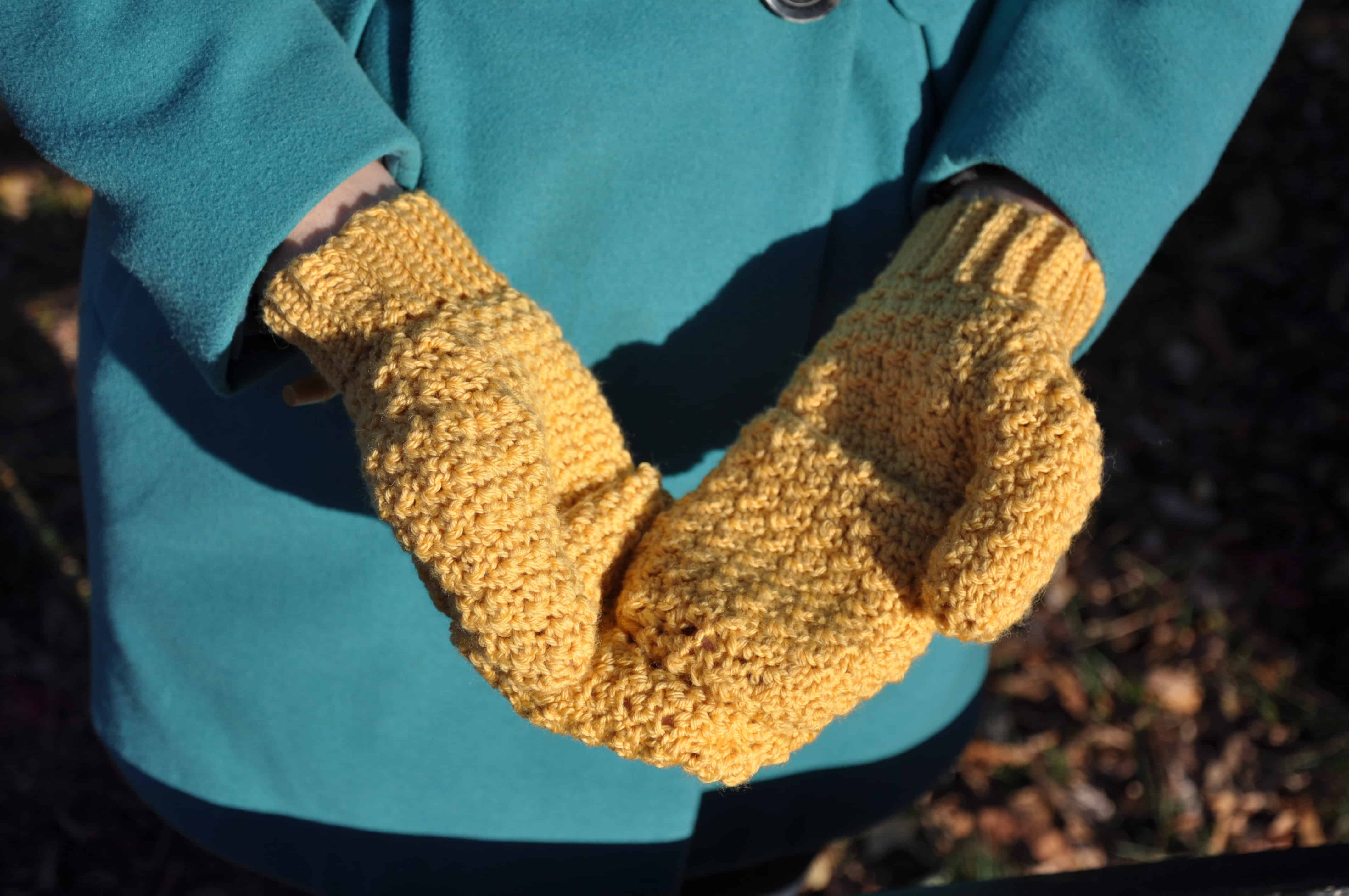 A person wearing a teal coat holds their hands together, palms up, while wearing yellow crochet mitts outdoors.