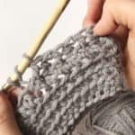 A person is crocheting a grey stitch using a crochet hook.