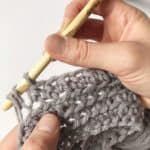 A person is using a crochet hook to knit a grey sweater pattern.