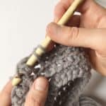 A person using a crochet hook to knit a grey sweater with a pattern.