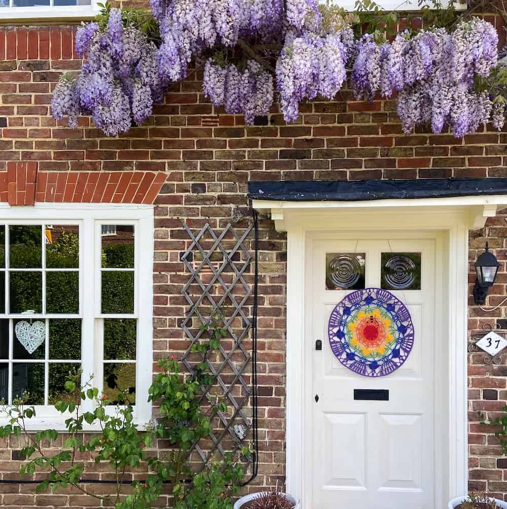 a rainbow crochet mandala hanging on a period property front door with wisteria hanging over.