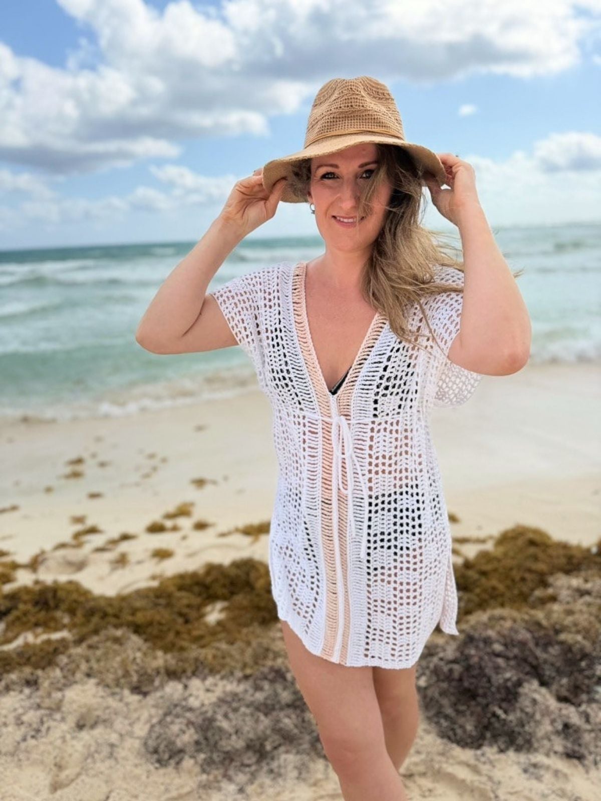 A person wearing a white bikini cover up crochet pattern and a straw hat stands on a sandy beach with seaweed, ocean waves, and a partly cloudy sky in the background.
