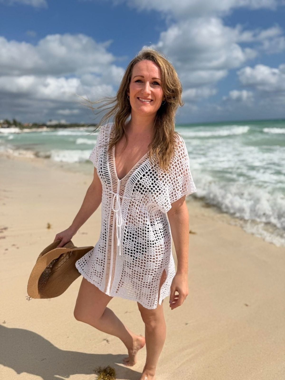 Woman in a white bikini cover up crochet pattern stands barefoot on sandy shore, holding a hat, with waves and partly cloudy sky in the background.