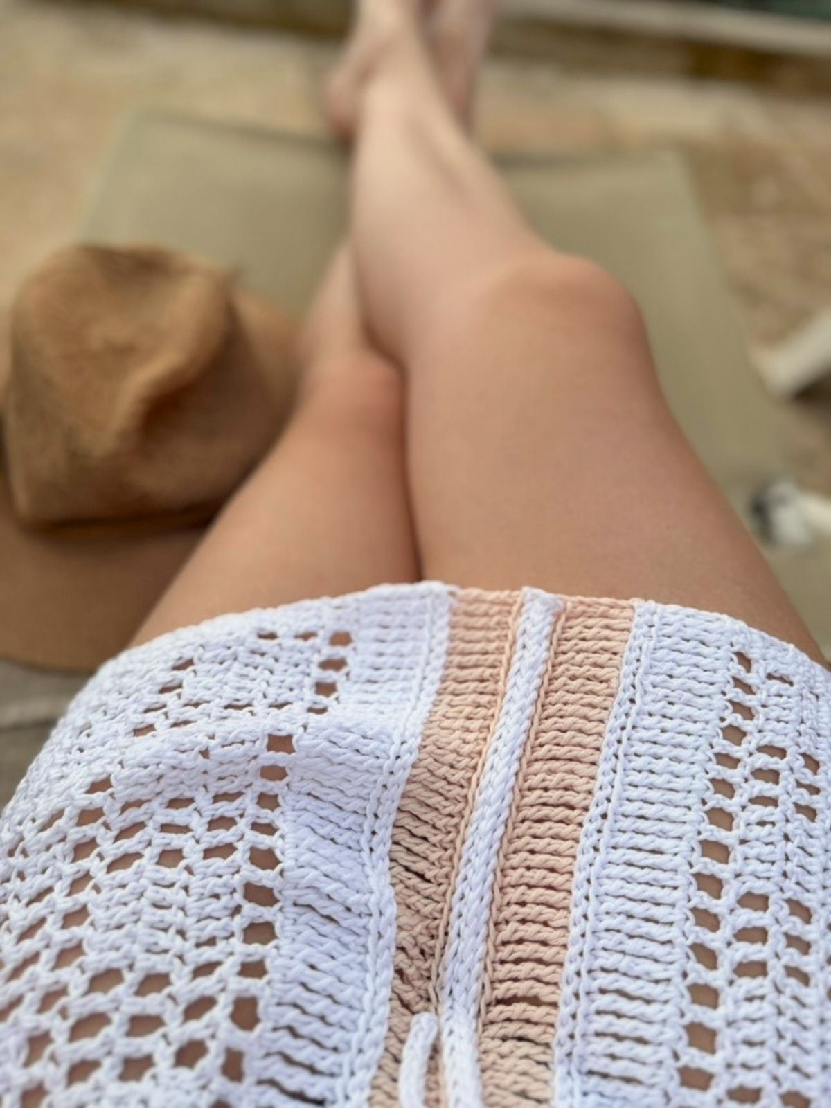 Person lounging on a chair, wearing a white bikini cover up crochet pattern, with a brown hat placed beside them. The background appears to be outdoors.