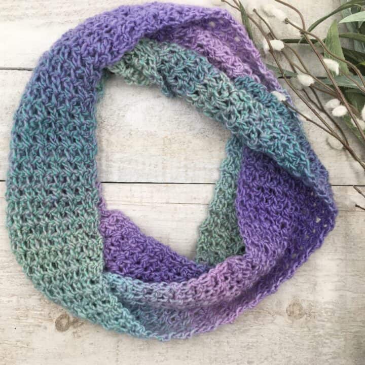 green and purple crochet infinity scarf laying on white floor with leaves