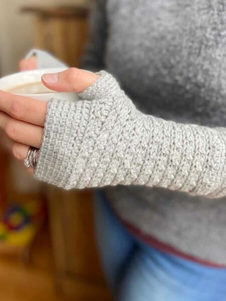 woman holding cup of coffee wearing grey fingerless crochet mittens