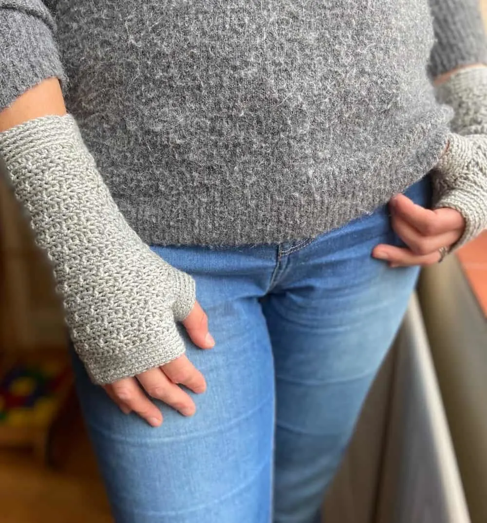 Woman in jeans with hand in pocket wearing grey fingerless mittens.