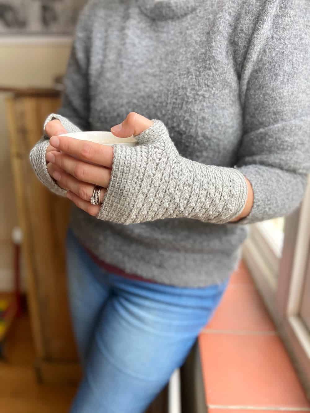 Woman leaning against windowsill wearing grey fingerless crochet mittens.