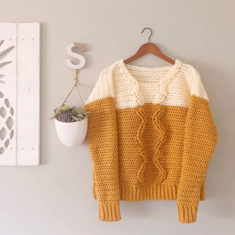 mustard and cream crochet cable sweater on hanger against grey wall with potted plant