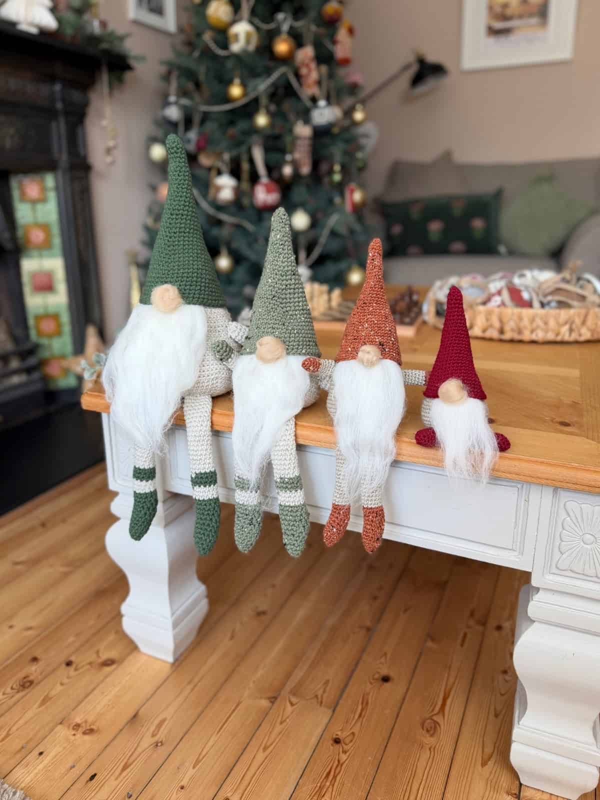 Four knitted gnome dolls with white beards and pointed hats sit on the edge of a wooden table in a Christmas-decorated living room, perfect inspiration if you're searching for a free crochet gnome pattern.