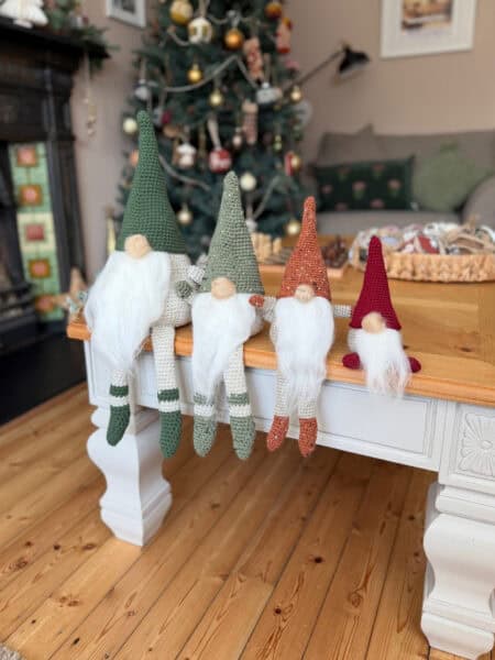 Four crochet gnome dolls with long white beards and pointed hats sit on a wooden table in a living room with a decorated Christmas tree—perfect inspiration if you’re looking for a free crochet gnome pattern this holiday season.