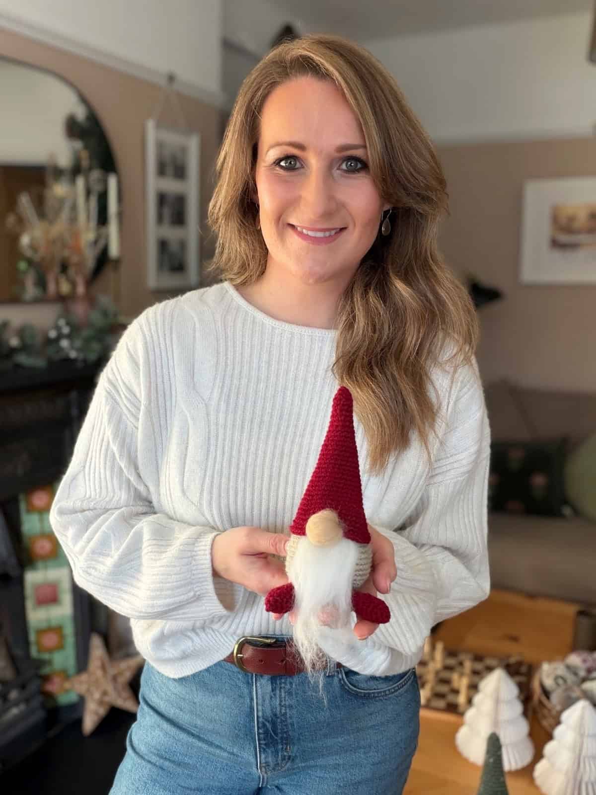 A woman in a white sweater and jeans holds a small gnome with a red hat, made from a free crochet gnome pattern, standing in a cozy living room decorated for the holidays.