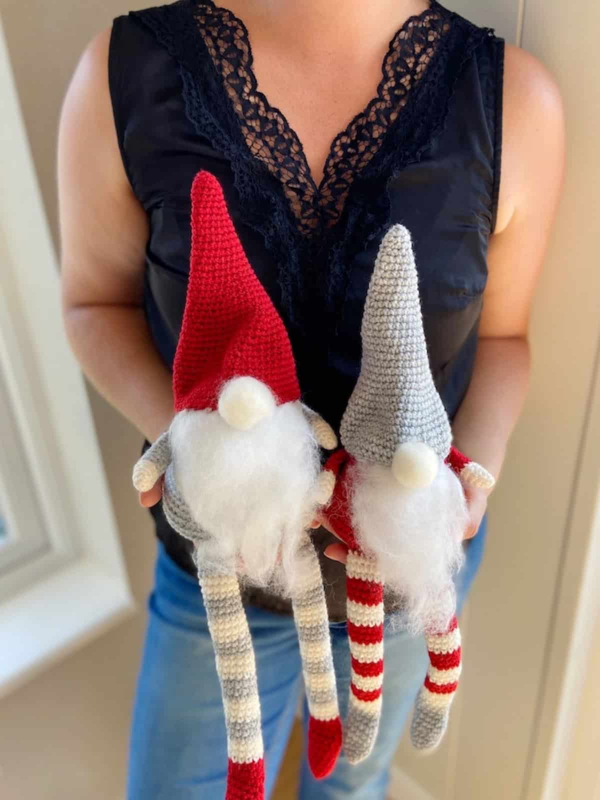 A person in a black lace-trimmed top holds two knitted gnomes with striped legs, white beards, and pointed hats—one red, one gray—made from a free crochet gnome pattern.