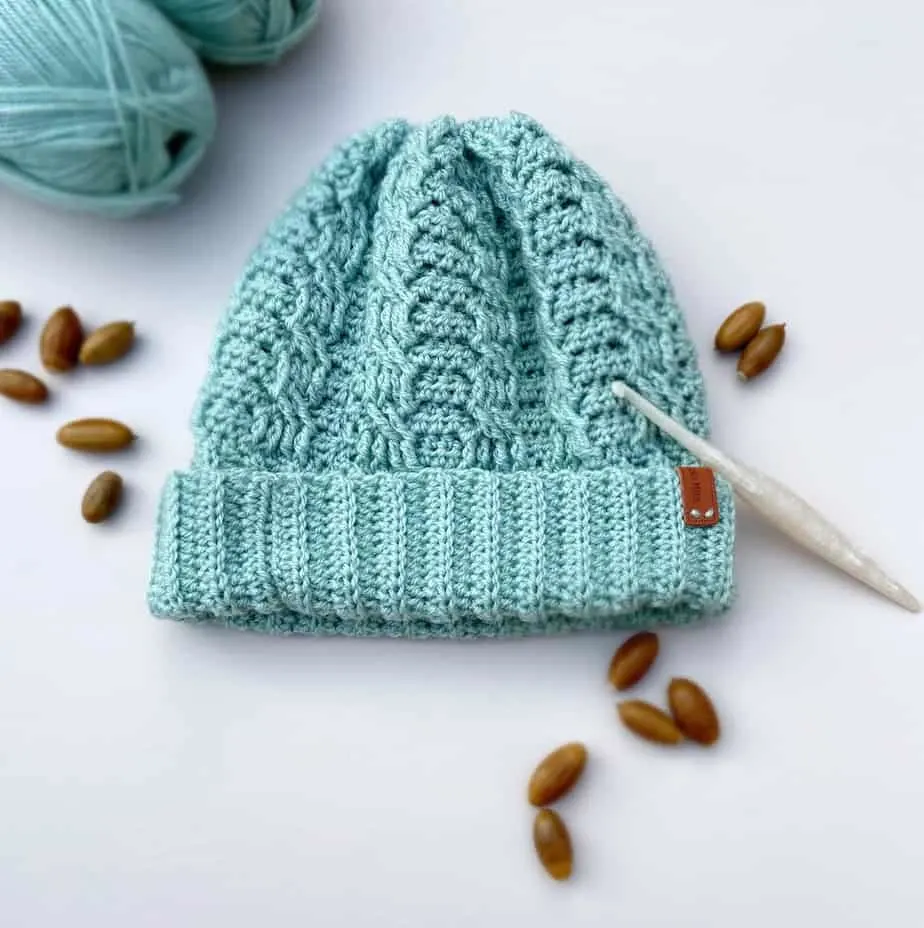 mint green crochet cable hat with wide brim laid on table with crochet hook and acorns