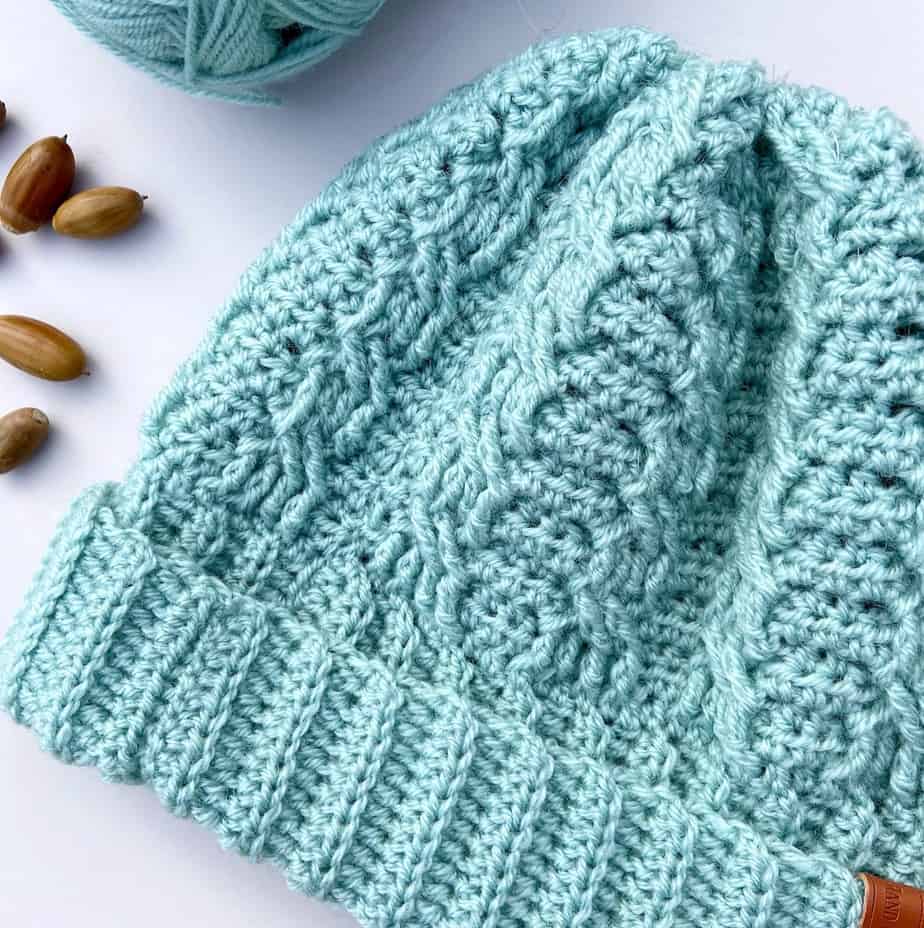 close up of mint green crochet cable hat with crochet hook and acorns laying on table