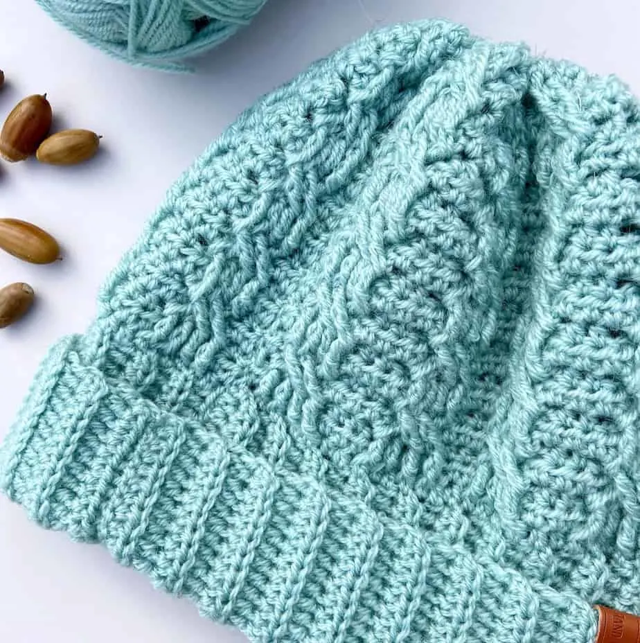 Close-up of mint green crochet cable hat with crochet hook and acorns laying on table.