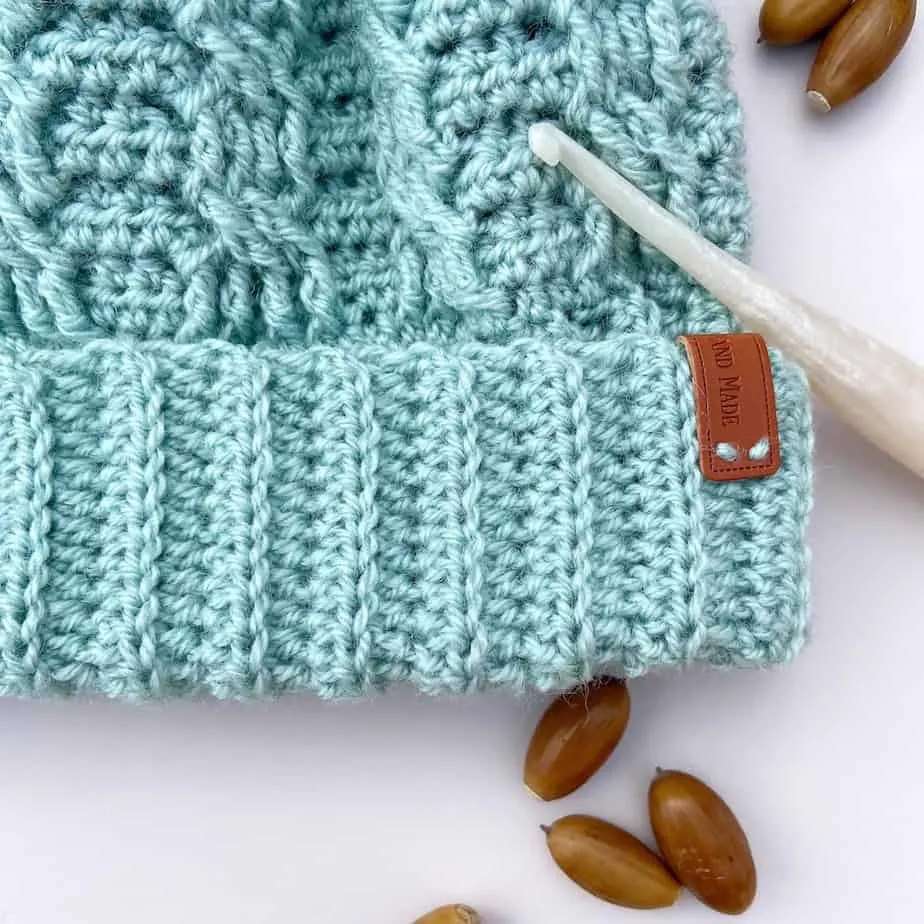 Close up of mint green crochet cable hat with crochet hook and acorns laying on table.