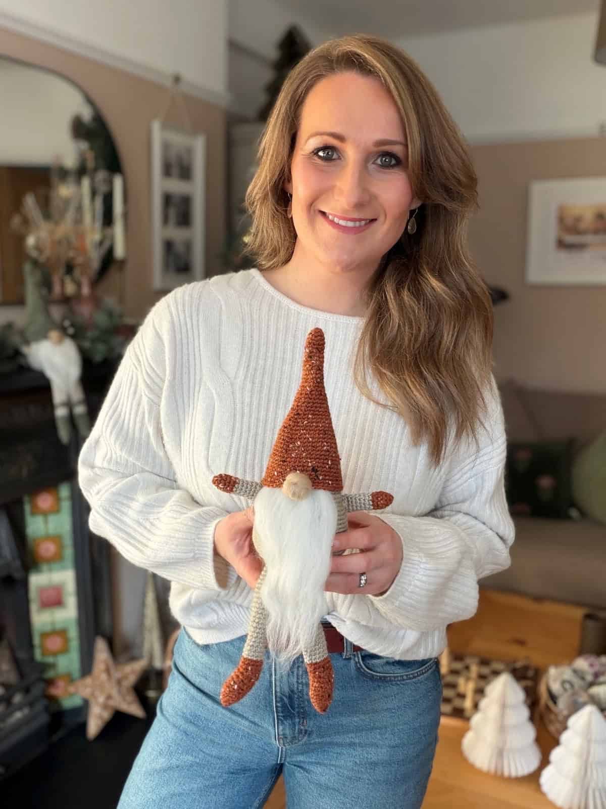 A woman in a white sweater and jeans stands indoors, smiling and holding a small decorative gnome with a pointed hat and white beard—crafted from a free crochet gnome pattern.