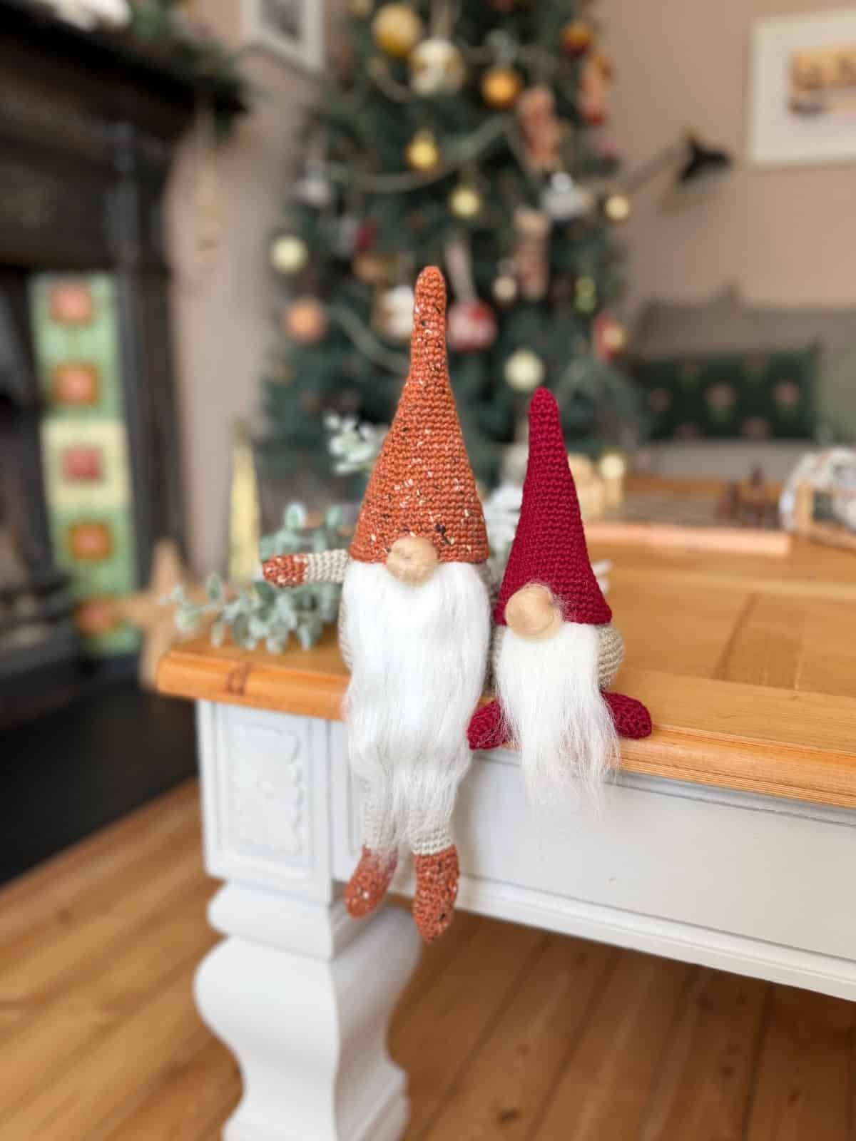 Two knitted gnome dolls with white beards and pointed hats sit on the edge of a wooden table; a decorated Christmas tree is in the background—perfect inspiration for your next project using a free crochet gnome pattern.