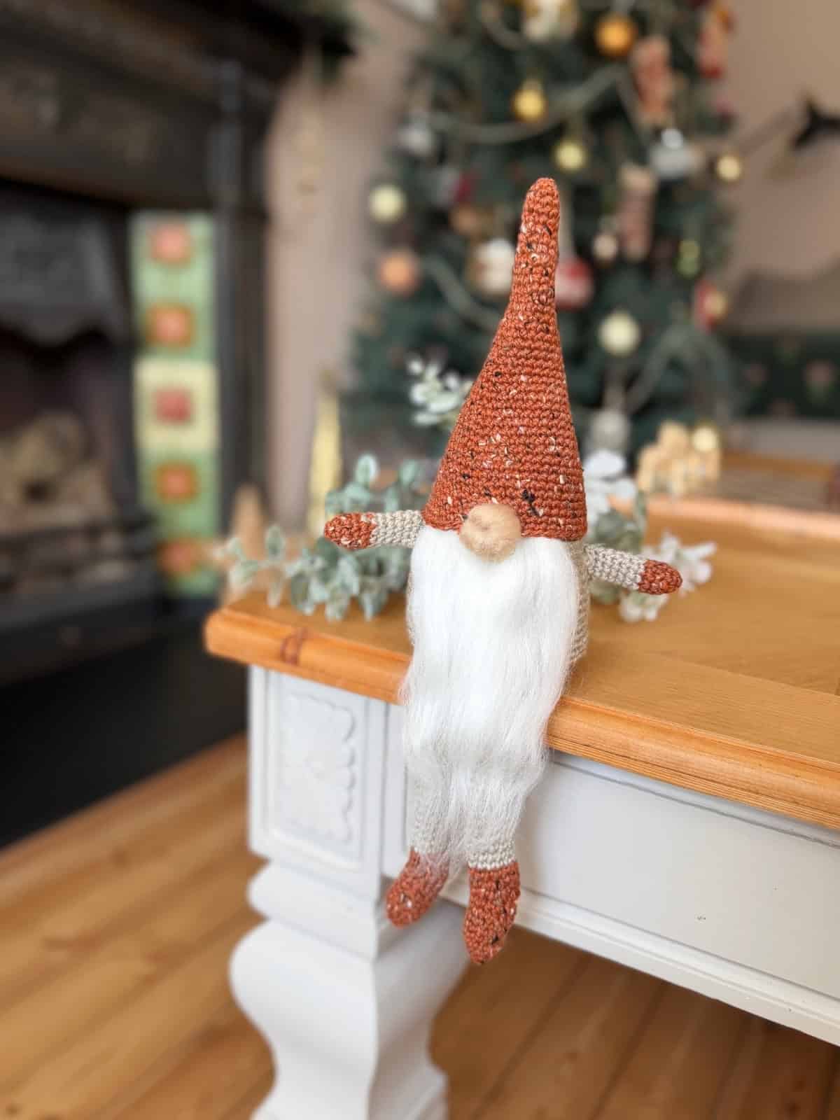 A crochet gnome with a tall red hat and white beard sits on the edge of a wooden table near a decorated Christmas tree, inspiring you to try your own free crochet gnome pattern this holiday season.