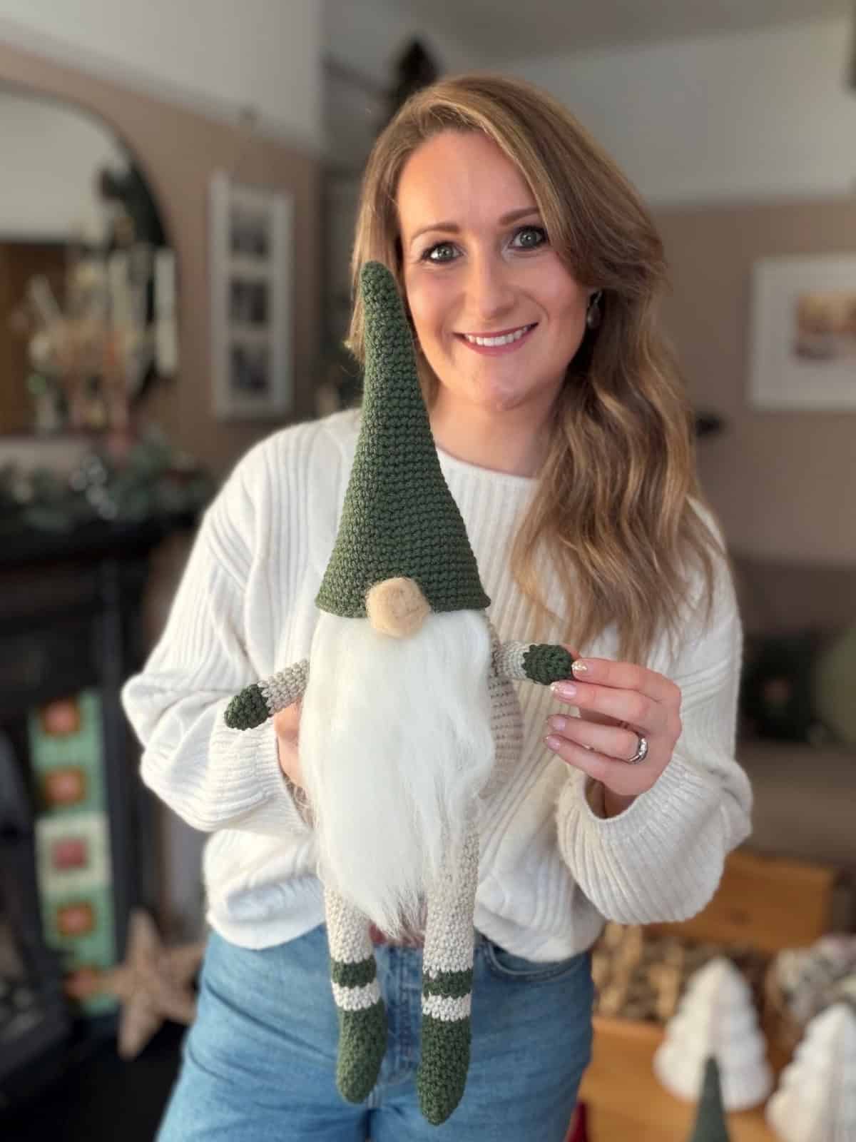 Woman in a white sweater and jeans holding a knitted gnome doll with a green hat and white beard, standing in a cozy, decorated living room—perfect inspiration if you're looking for a free crochet gnome pattern.