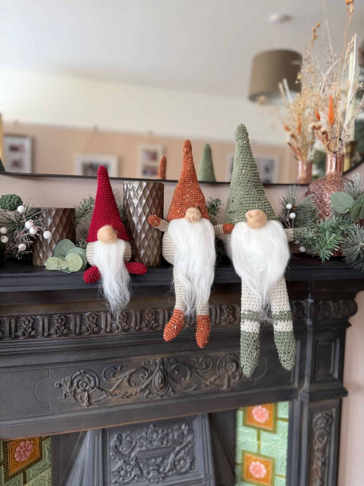 Three knitted gnome decorations with long white beards sit on a decorated mantelpiece with greenery and vases in the background, perfect for those inspired by a free crochet gnome pattern.