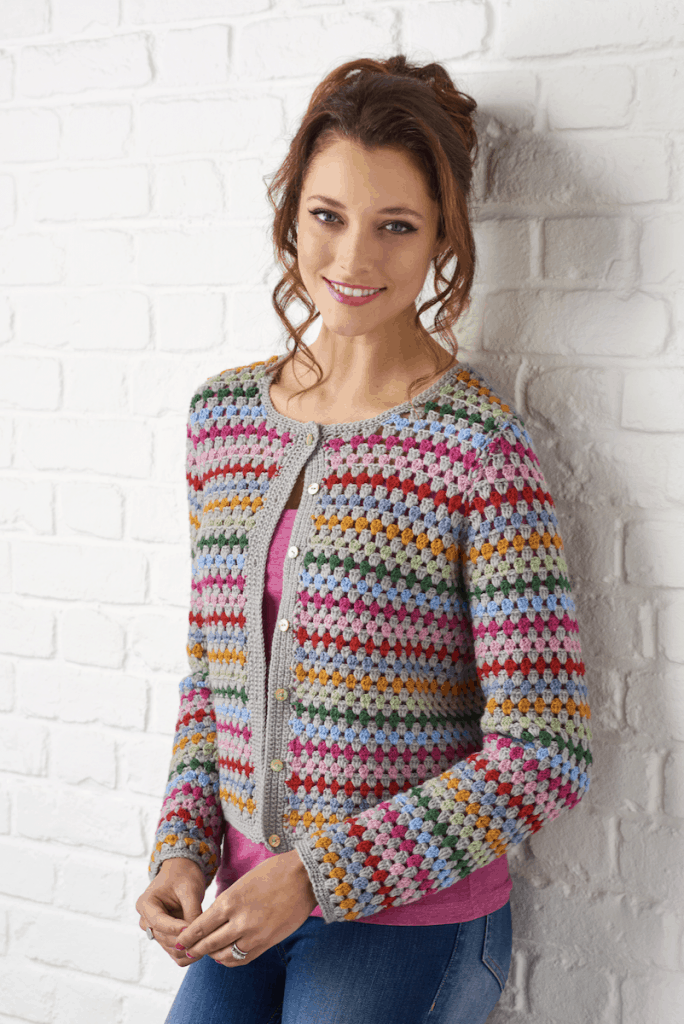 Woman leaning against wall wearing colourful granny stripe crochet cardigan and jeans.