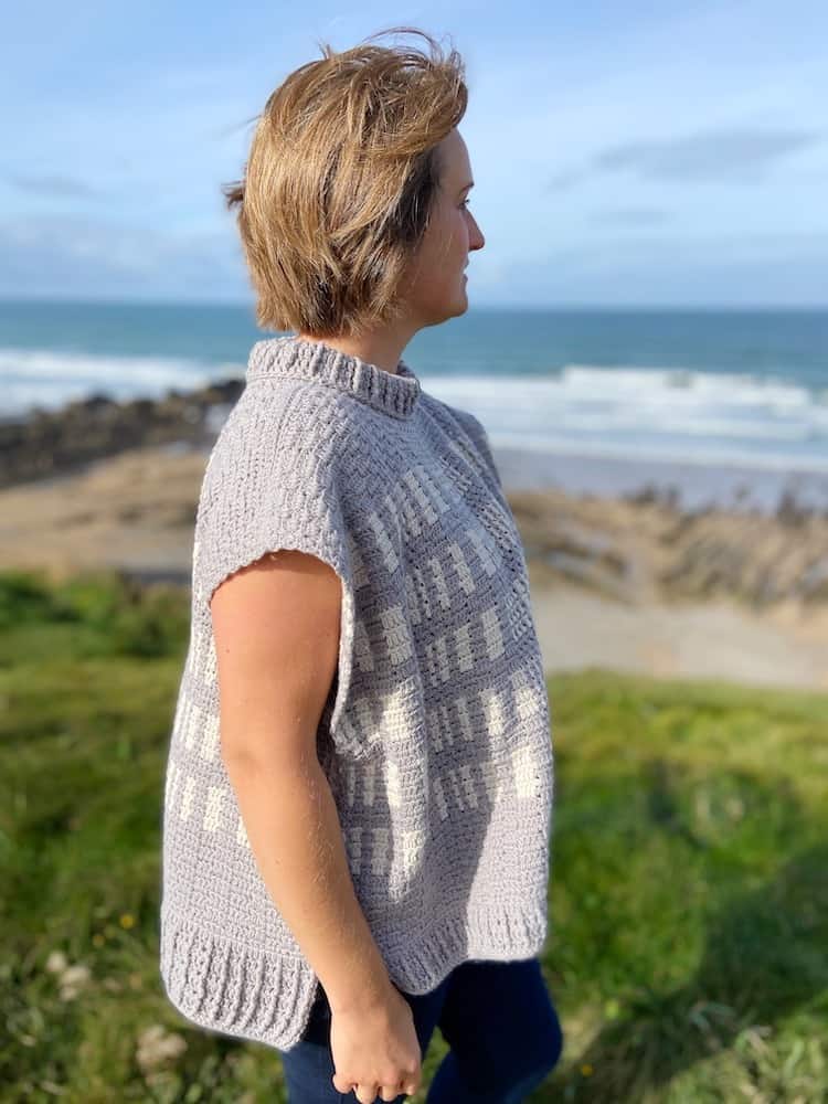 woman standing on cliff top wearing grey and white crochet poncho with hair blowing in the wind