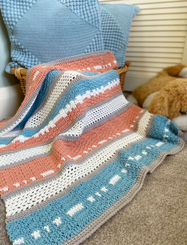 Modern peach and teal crochet baby blanket draped over basket with cushions and cuddly toy.