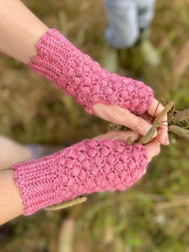 close up of hands wearing pink puff stitch crochet mittens holding grass in a field with little boy playing in the background