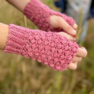 close up of hands wearing pink puff stitch crochet mittens with little boy playing in the background