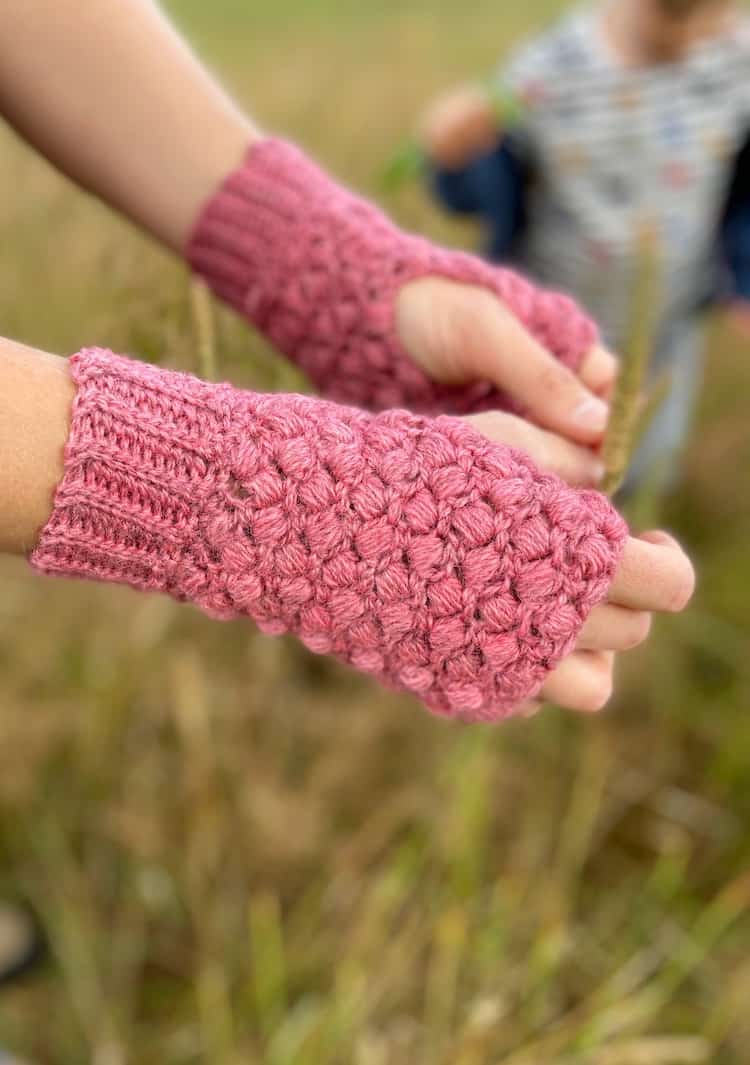 close up of hands wearing pink puff stitch crochet mittens with little boy playing in the background