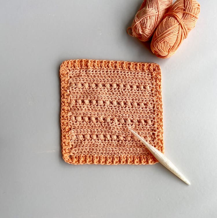 flatlay image of peach crochet puff stitch square design with yarn and Furls crochet hook.