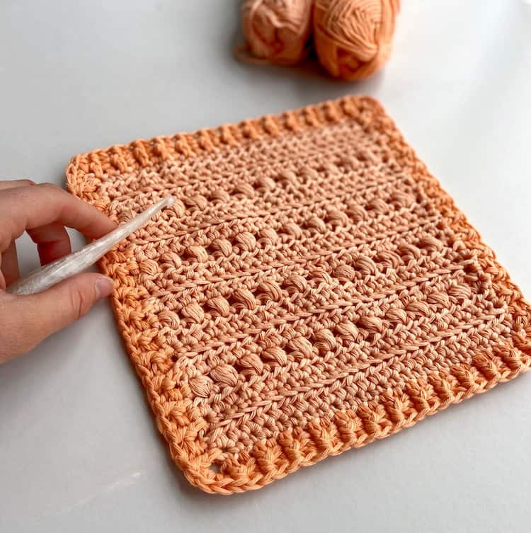 peach crochet puff stitch square motif on table with hand holding crochet hook and yarn in background.