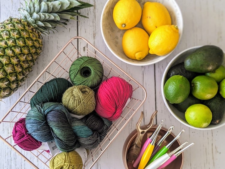 basket of pink and green yarn with crochet hooks surrounded by bowls of lemons, limes and a pineapple