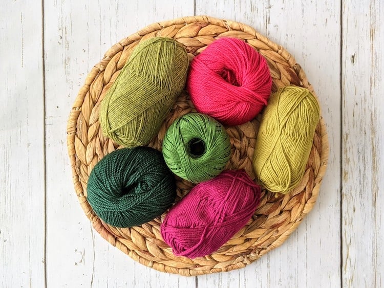 6 balls of pink and green yarn sitting on woven mat on table