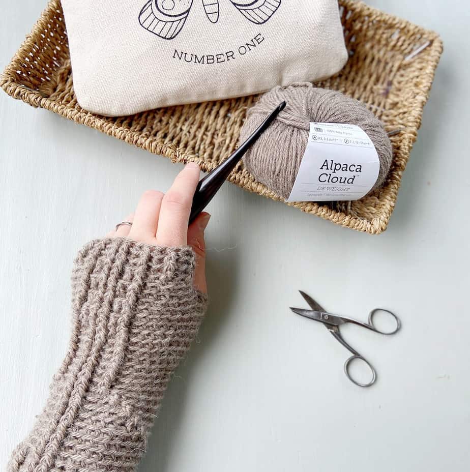 left hand wearing crochet mitten holding a crochet hook with scissors, yarn and canvas pouch on desk