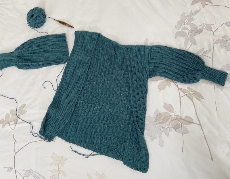 a knit-look crochet cardigan laid flat on a bed with crochet hook and yarn, one sleeve hasn't been joined yet.