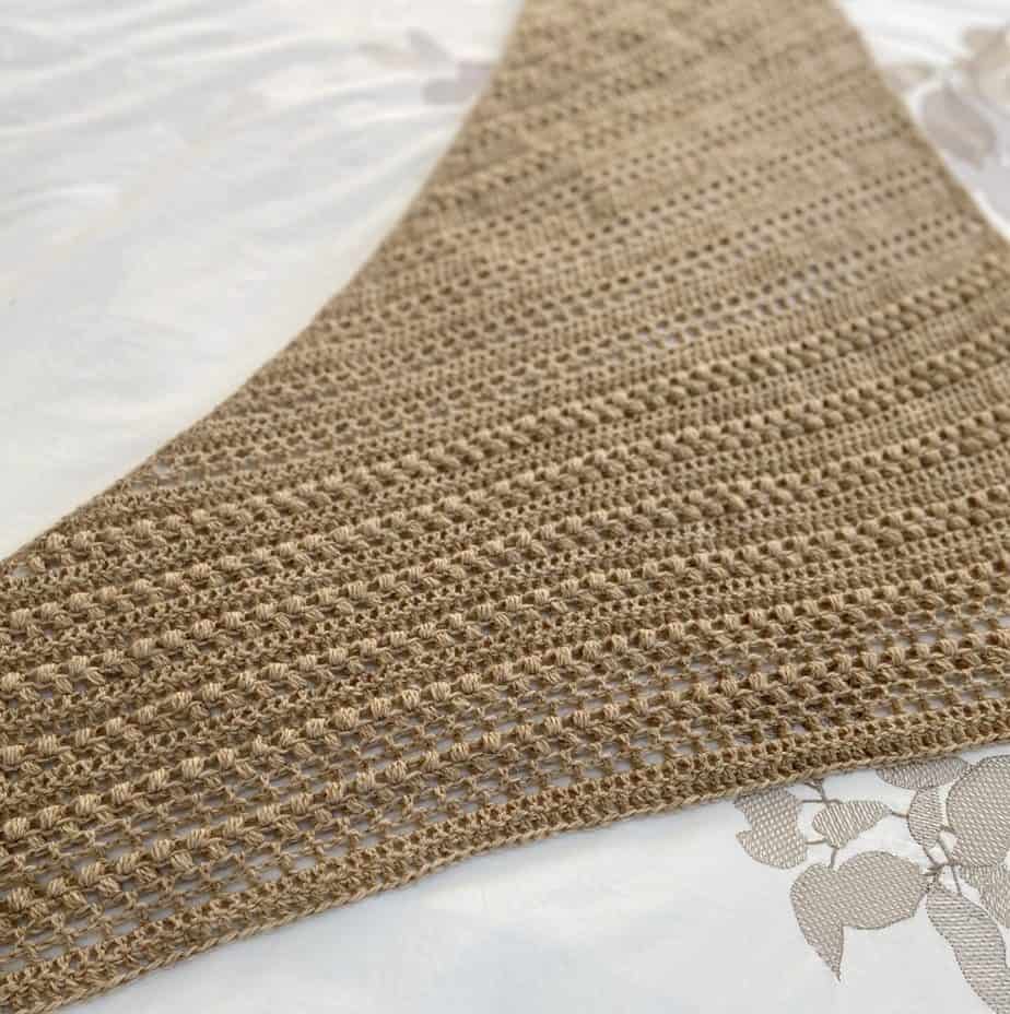 a textured, beige triangular crochet shawl laid flat on a white bed sheet