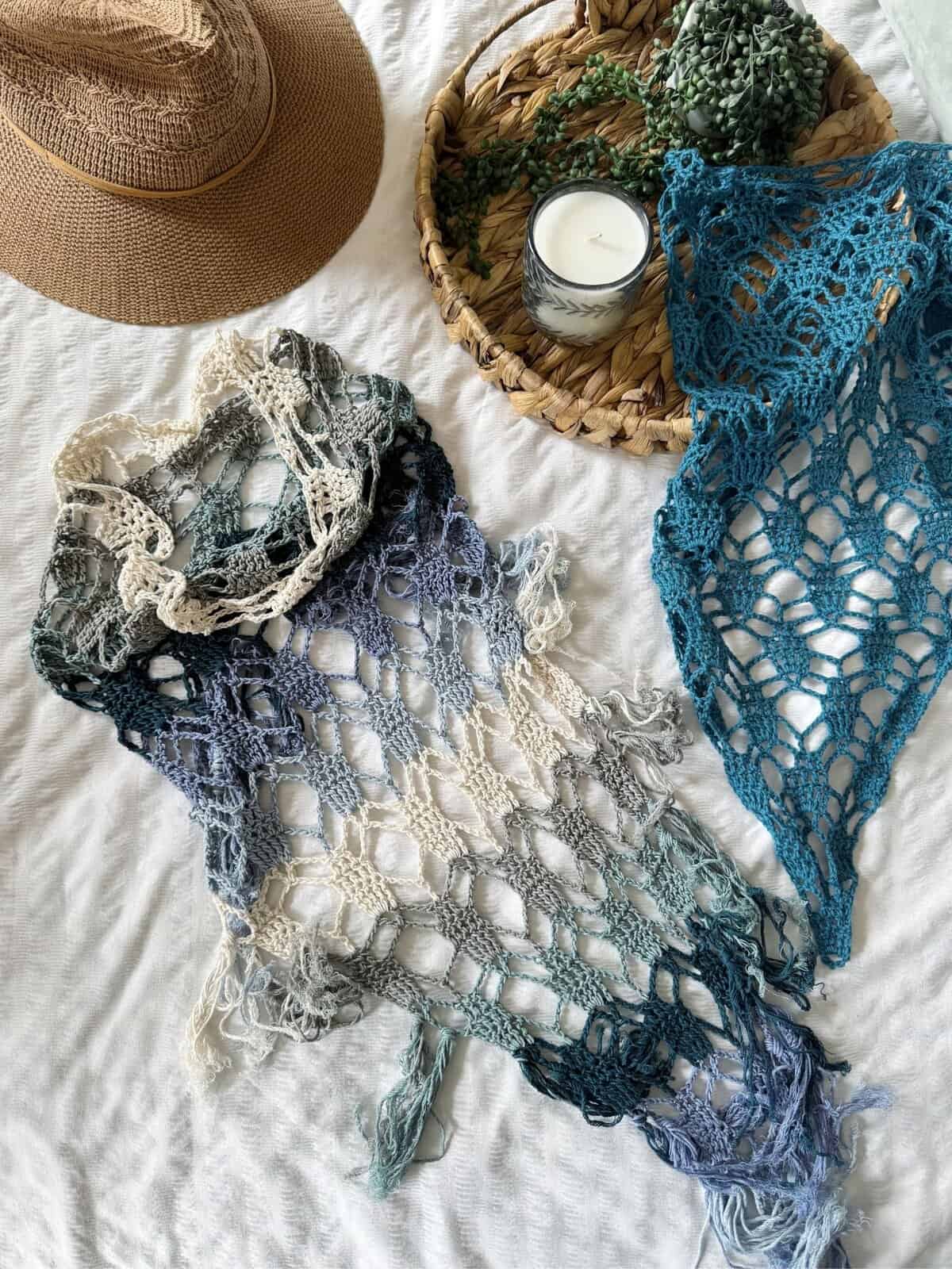 A straw hat, a woven tray with a candle and greenery, and two crochet boomerang shawl scarves in blue and cream tones are arranged on a white textured surface.