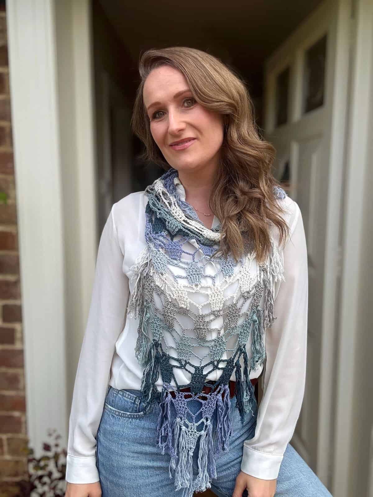 A woman with wavy brown hair stands indoors, wearing a white blouse, blue jeans, and a vibrant crochet boomerang shawl with fringe.