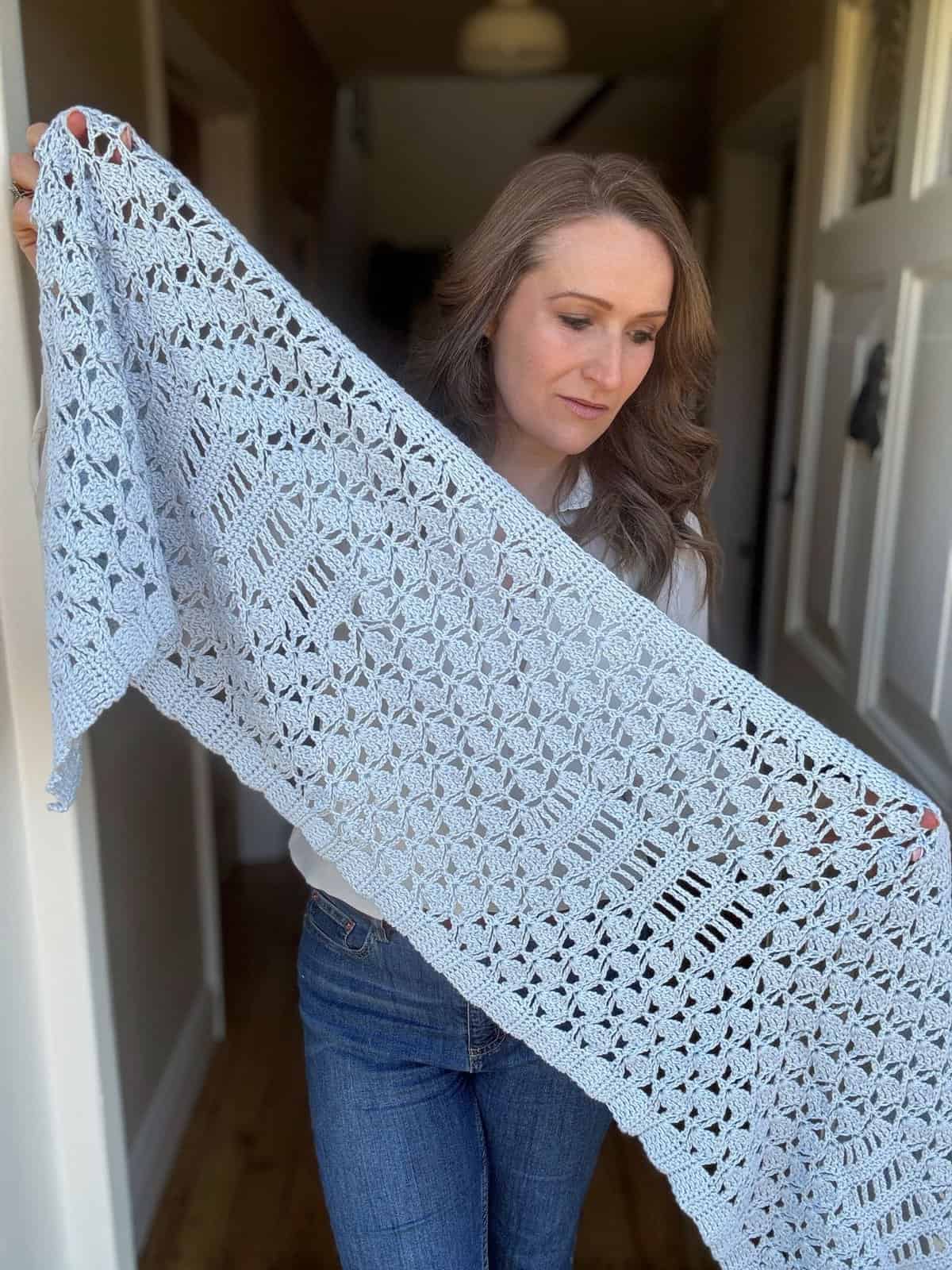 A woman stands indoors holding and displaying a light blue crochet boomerang shawl with a striking geometric lace pattern.