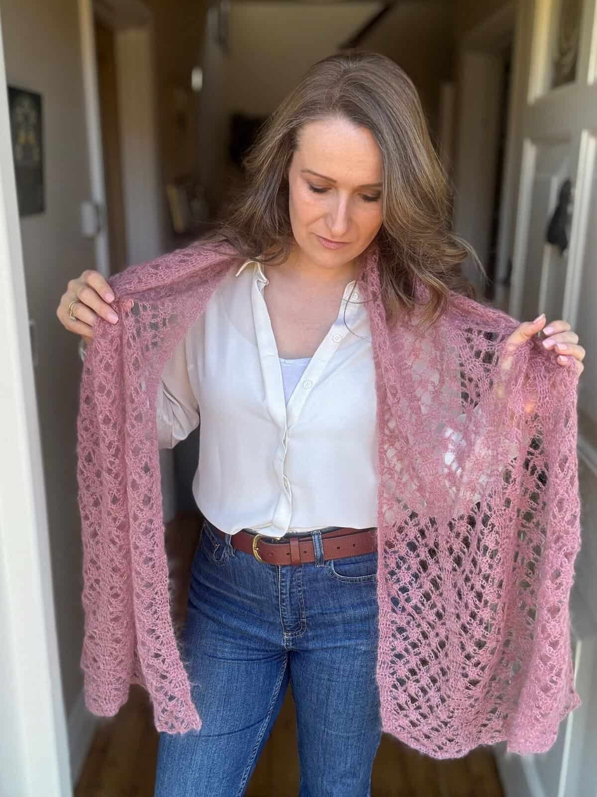 A woman stands indoors, looking down, holding out a pink, lacy crochet boomerang shawl with both hands. She is wearing a white blouse and blue jeans.