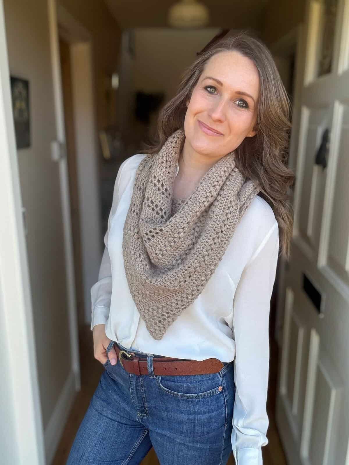 Woman standing indoors, wearing a white blouse, blue jeans, brown belt, and a beige crochet boomerang shawl as a scarf, smiling slightly while looking at the camera.